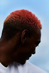 Profile portrait of a Black man with short red dyed hair wearing a white t shirt against a clear sky background