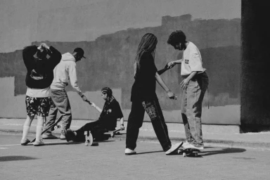 Monochrome street-level photograph of five people skating and interacting outdoors against a textured wall.