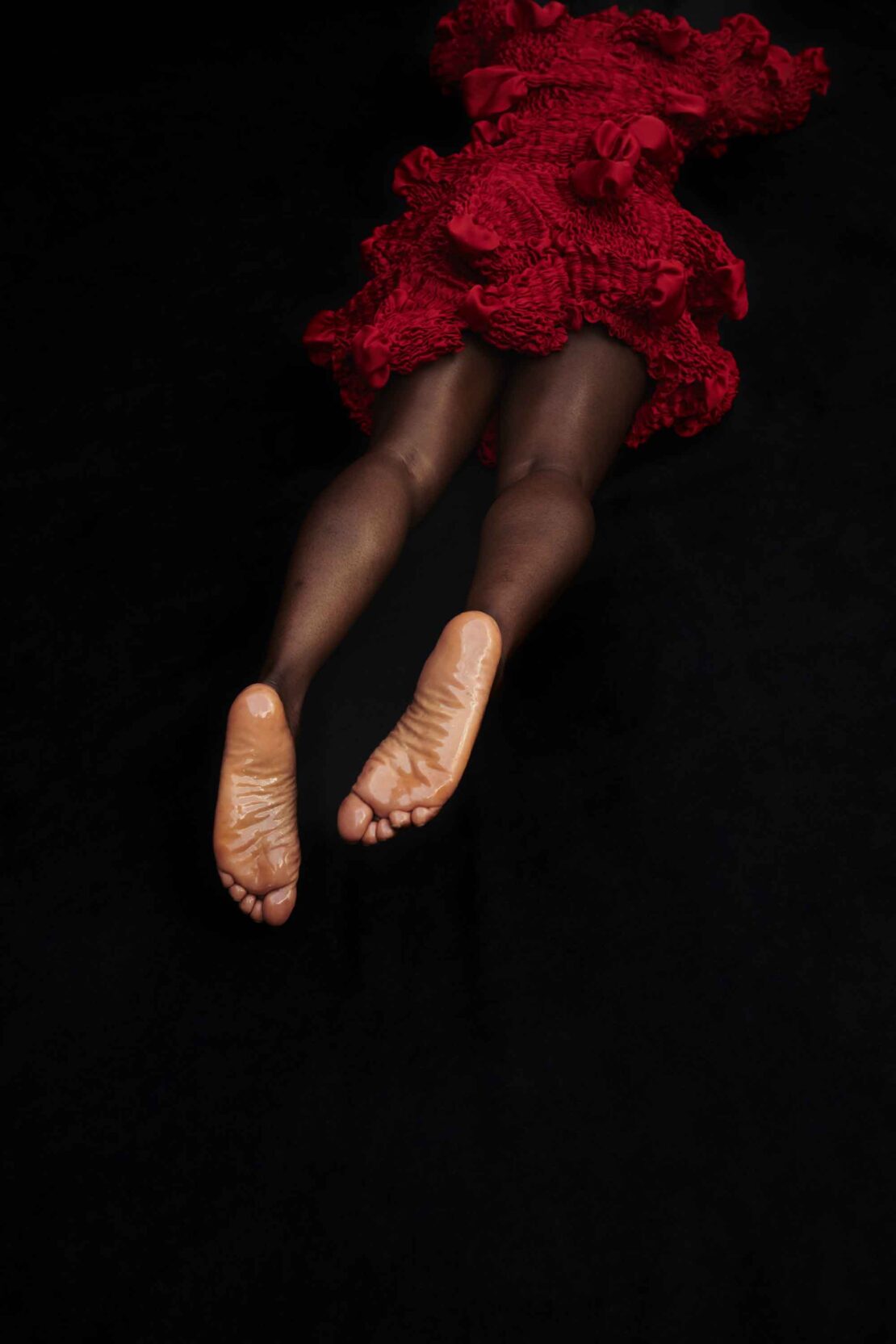 Back view of a person in a red, textured dress with visible legs and feet against a dark background, emphasizing dark skin tones.