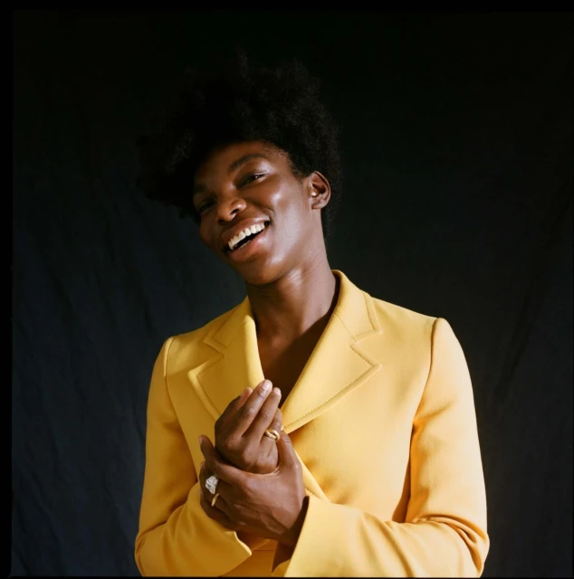 Smiling Black woman in yellow blazer poses indoors against dark background, showcasing vibrant fashion and joyful expression.
