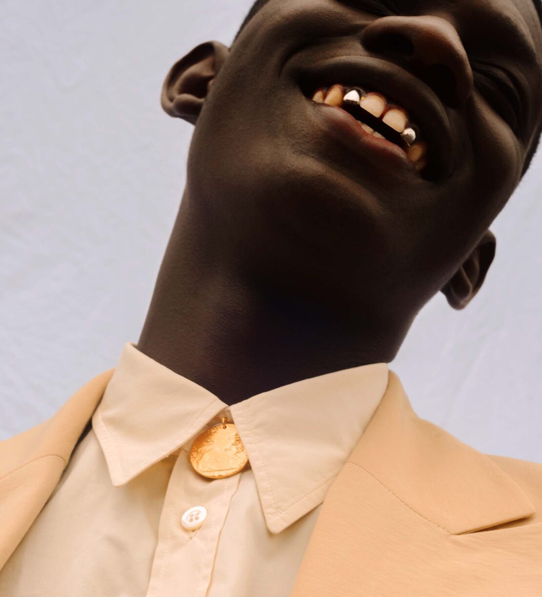 Close-up of a smiling Black man wearing a beige suit with a gold necklace, showcasing a fashion or portrait aesthetic.
