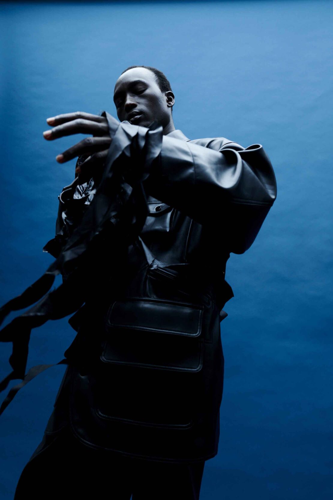 A person in a dark jacket and a dark shirt poses against a blue background, creating a strong contrast of light and shadow in a studio setting.