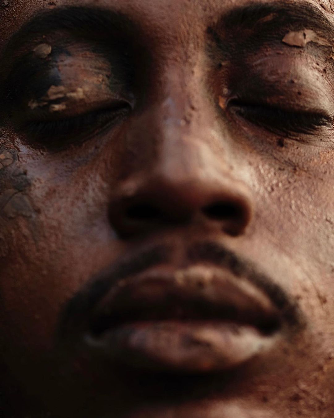 Close-up macro portrait of a Black man's face covered in wet reddish-brown mud, eyes closed.
