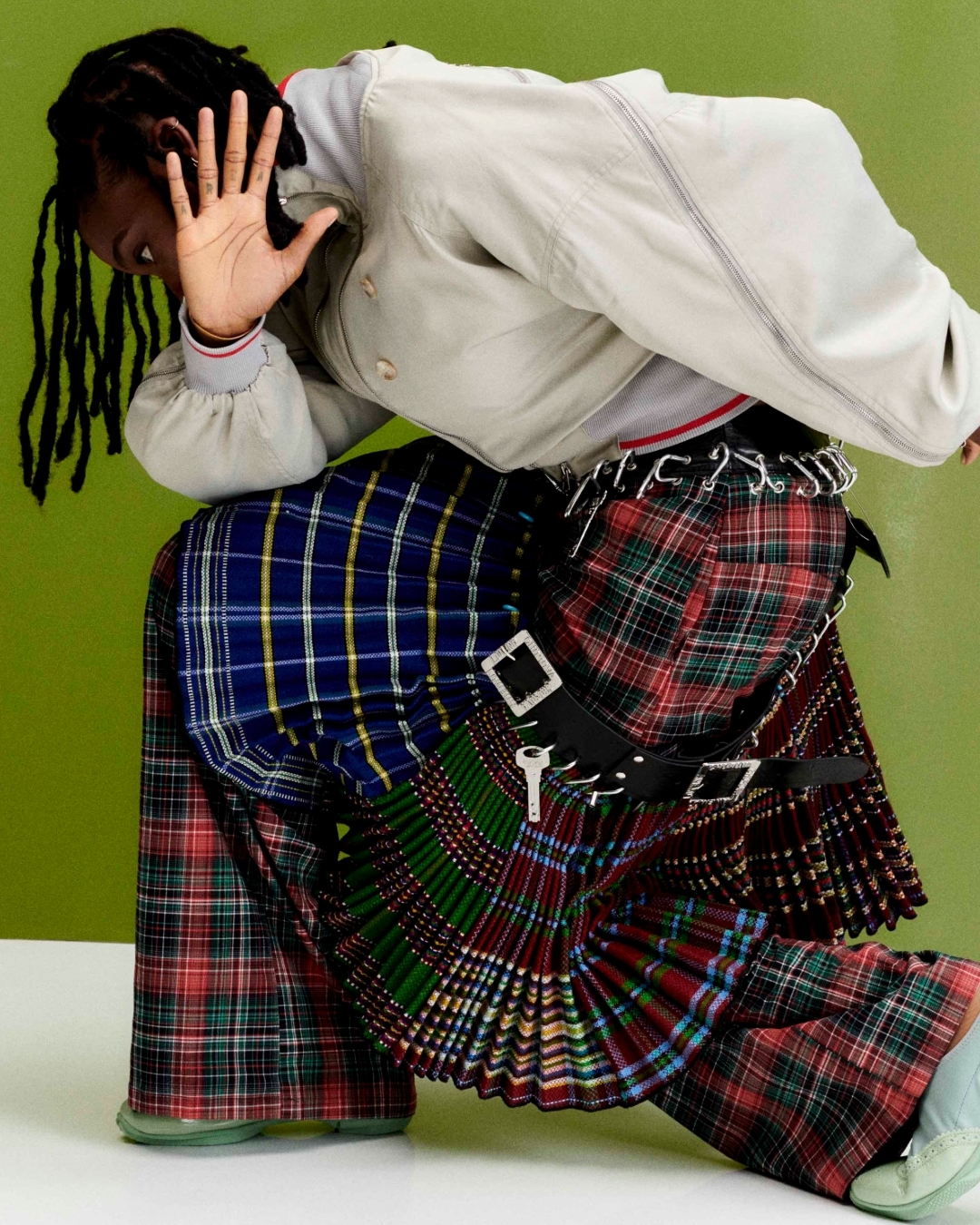 A person with dreadlocks, wearing a gray jacket and a multicolored plaid kilt, with a green wall in the background.