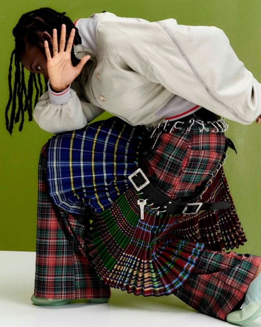 A person with dreadlocks, wearing a gray jacket and a multicolored plaid kilt, with a green wall in the background.