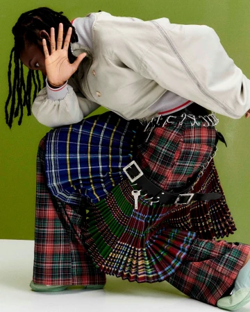 A person with dreadlocks, wearing a gray jacket and a multicolored plaid kilt, with a green wall in the background.