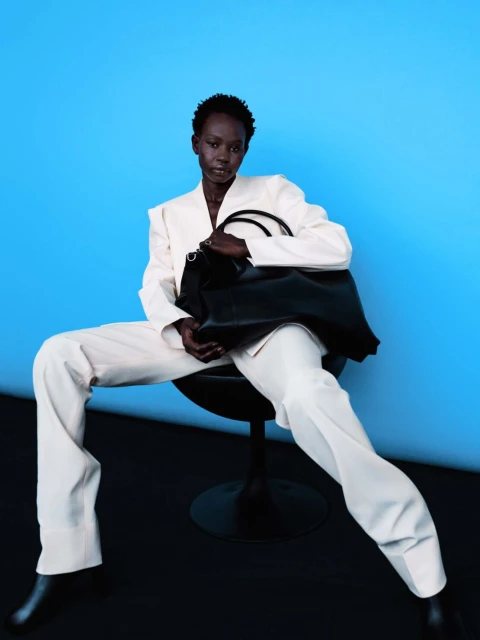 Dark-skinned model in a light suit poses on a black chair against a blue background, holding a large black leather tote bag.