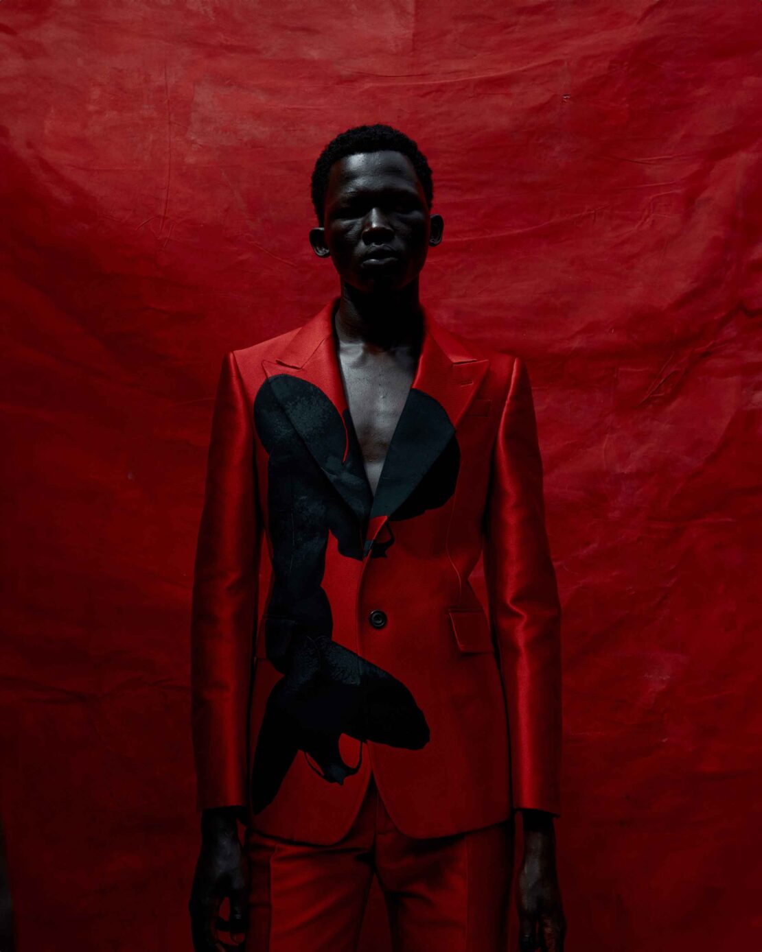 An adult Black male wearing a red suit with black graphic stands against a red textured backdrop in studio lighting.