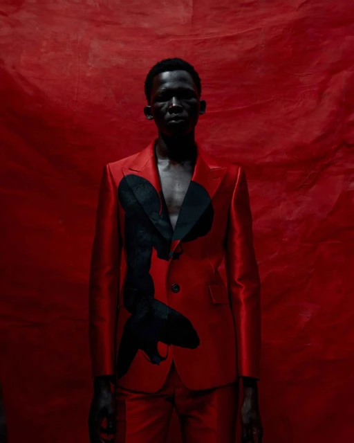 An adult Black male wearing a red suit with black graphic stands against a red textured backdrop in studio lighting.