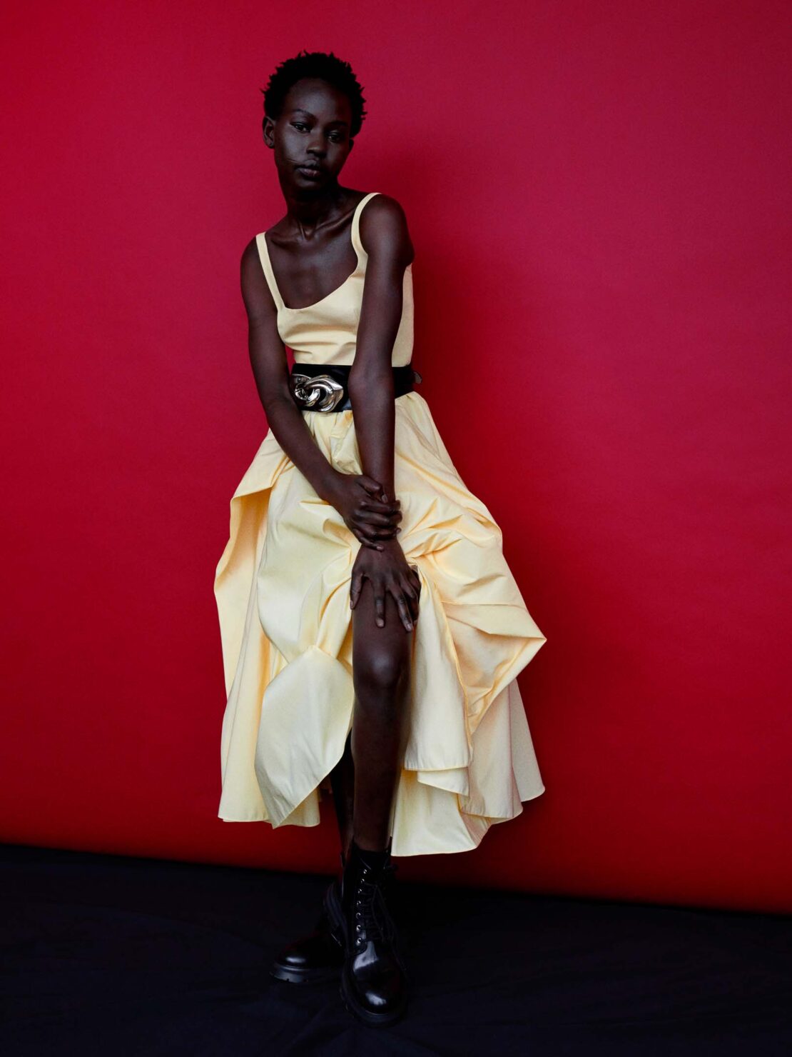 Fashion editorial shot of a Black model in a yellow dress with black boots against a red background, showcasing modern style and posing.