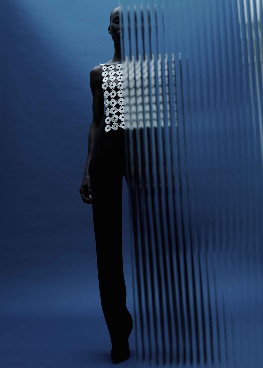 Artistic fashion shot featuring an adult woman in a white top and dark pants, posing behind a linear semi-transparent screen against a blue backdrop.