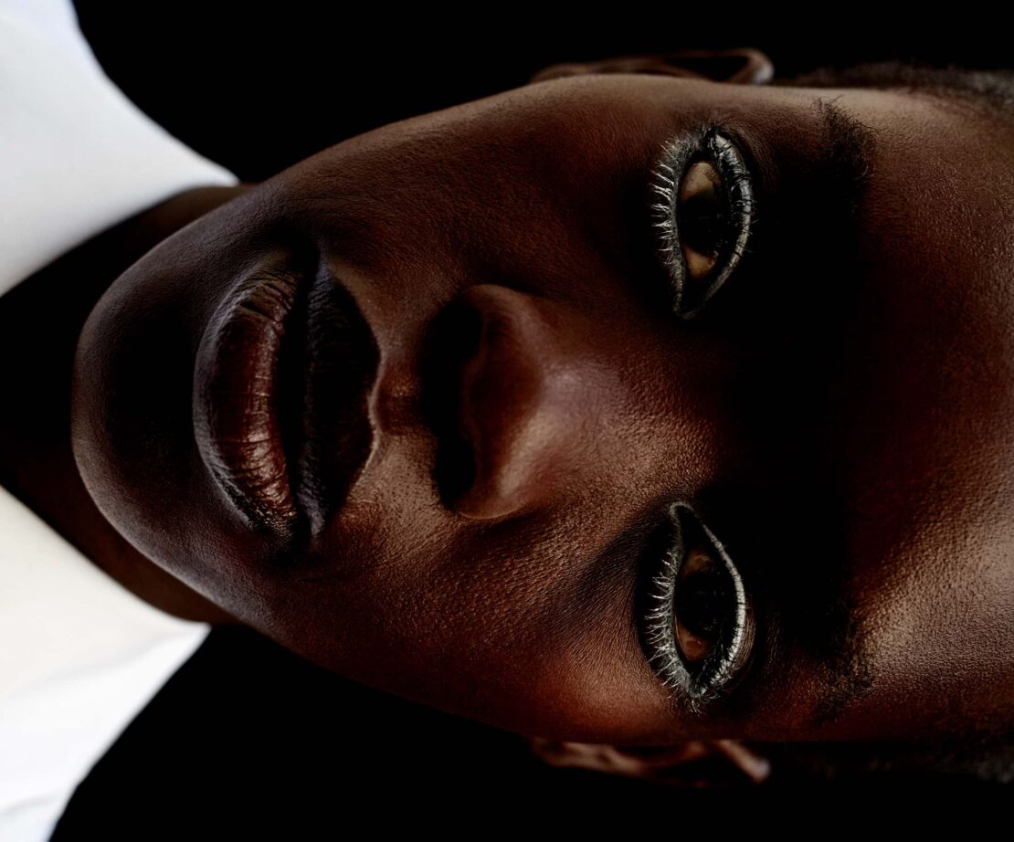 Close-up studio shot of a dark-skinned woman with glossy lips and dramatic eye makeup looking up against a dark background.