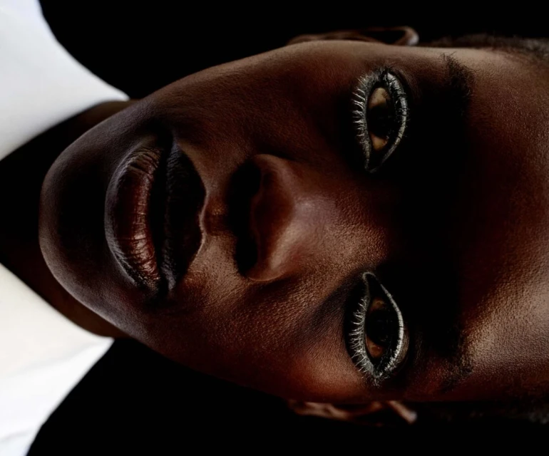 Close-up studio shot of a dark-skinned woman with glossy lips and dramatic eye makeup looking up against a dark background.