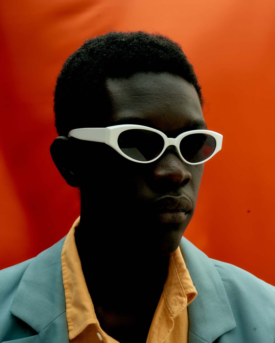 Portrait of a young man with dark skin wearing white sunglasses, a yellow shirt, and a light blue blazer against an orange backdrop.