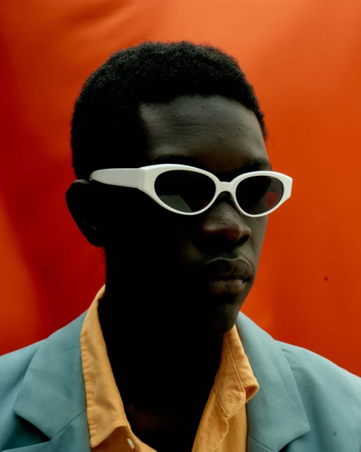 Portrait of a young man with dark skin wearing white sunglasses, a yellow shirt, and a light blue blazer against an orange backdrop.