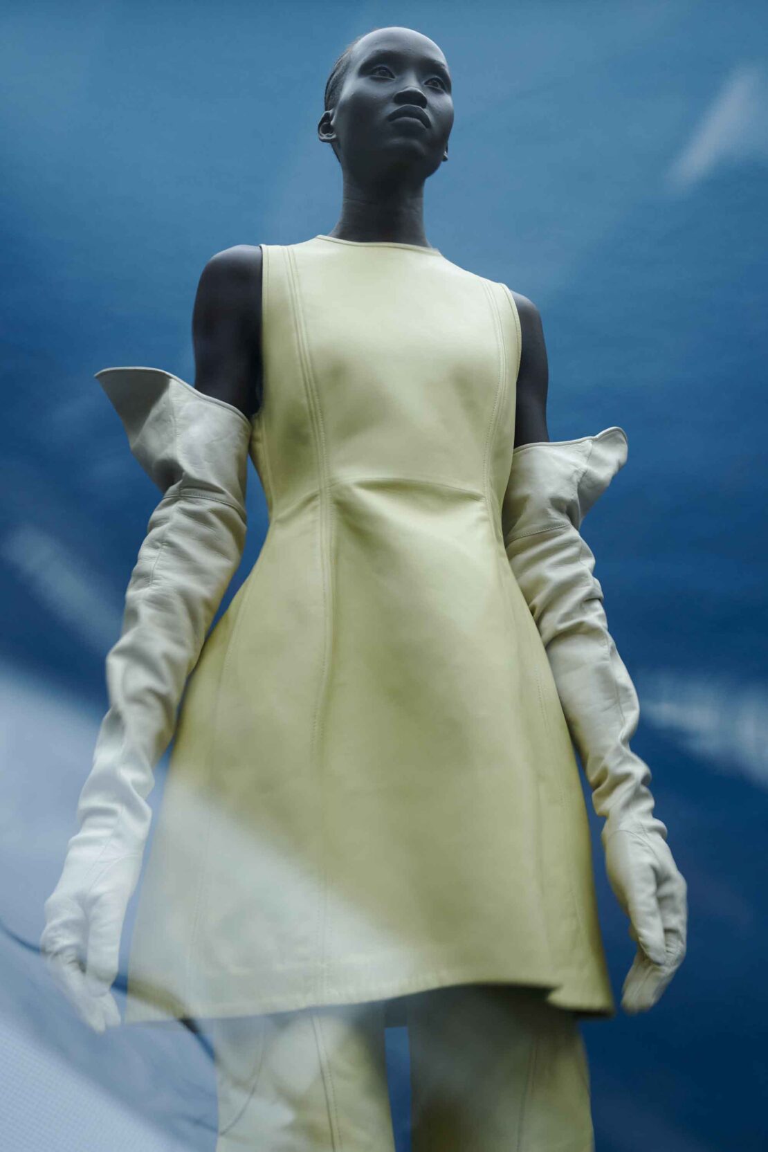 Woman in light yellow sleeveless mini dress and long white gloves against blurred blue background in fashion photography.
