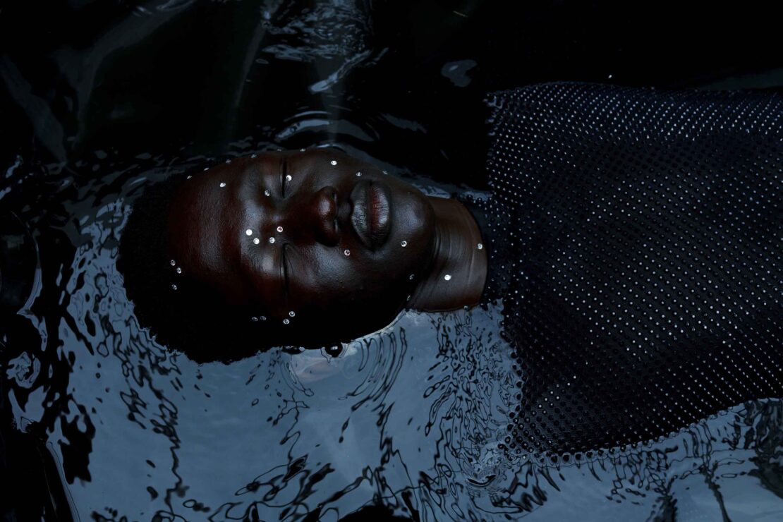 Black woman in water with reflective embellishments, dark and moody, artistic portrait featuring textured surface and water ripples.