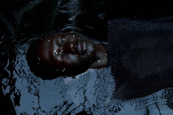 Black woman in water with reflective embellishments, dark and moody, artistic portrait featuring textured surface and water ripples.