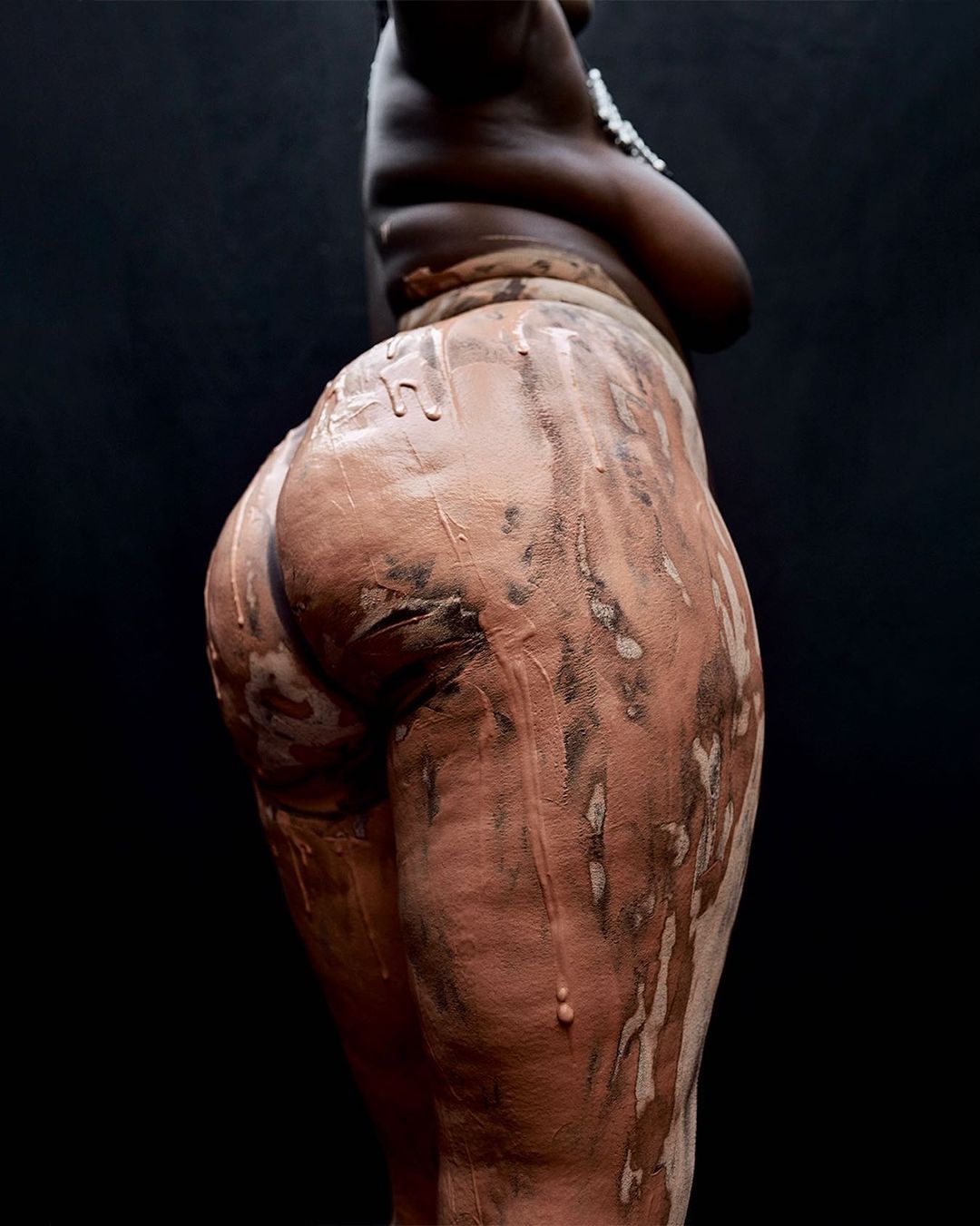 Striking studio shot of an African American woman's torso and legs, partially covered in clay, against a dark background, highlighting form and texture.
