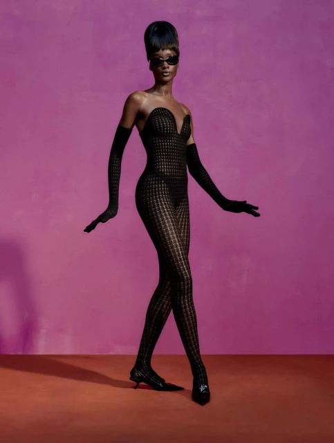 Fashion model in full body patterned sheer black bodysuit, long black gloves, sunglasses, and heels posing against a pink background.