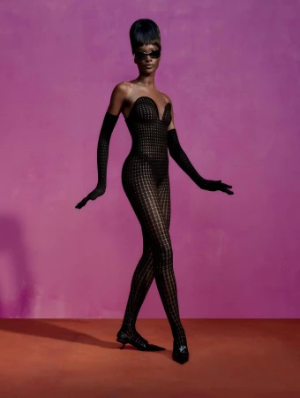 Fashion model in full body patterned sheer black bodysuit, long black gloves, sunglasses, and heels posing against a pink background.