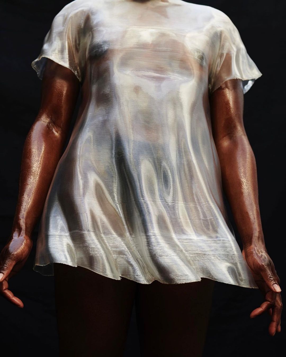 Close-up portrait of a Black person wearing a translucent glossy top against a dark black background, photographed by Ekua King.