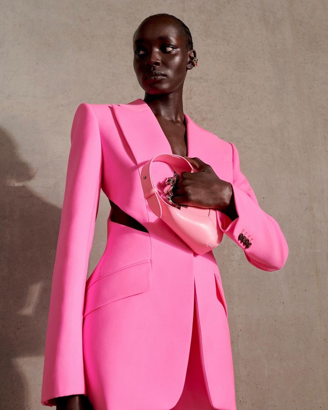 Model wearing a pink suit and holding a pink purse poses in front of a neutral wall in a studio setting showcasing fashion.