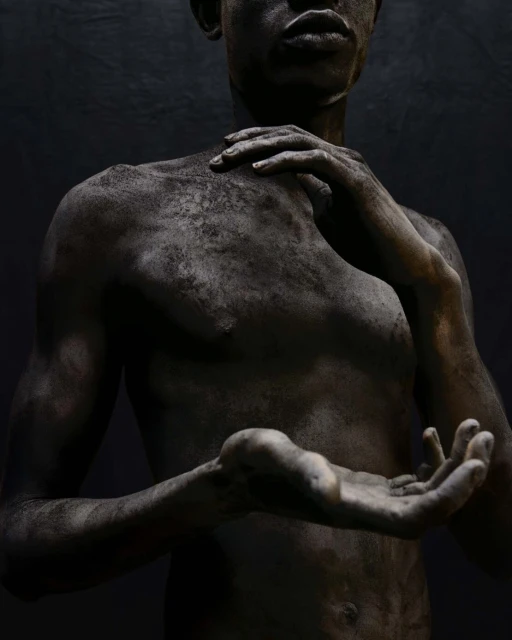 An adult human figure stands against a black background, the skin covered in a layer of dark particulate creating a dramatic, textured effect.