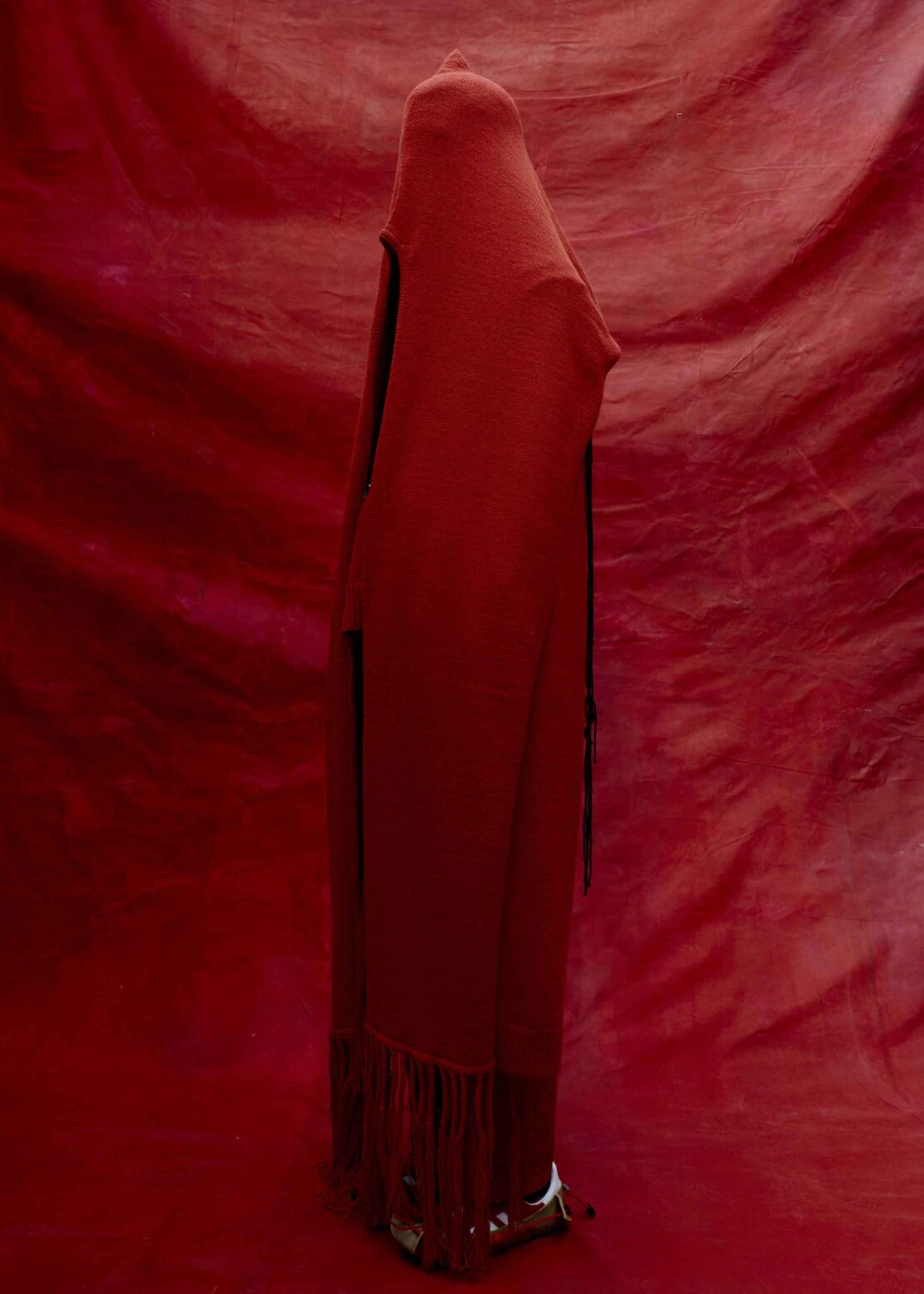 Person shrouded in a long red hooded garment stands against a red draped backdrop, creating a monochromatic and mysterious visual.