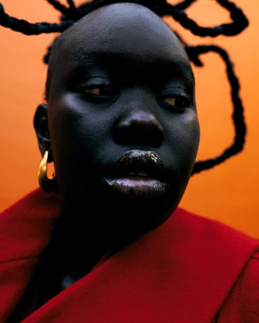 Close-up portrait of a Black woman with braided hair, gold hoop earrings, and glossy lips, against a bright orange background, and wearing a red jacket.