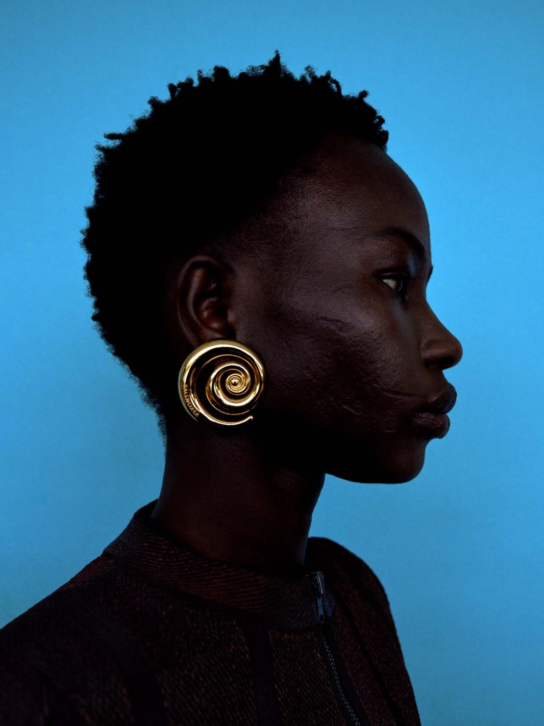 Profile portrait of a black woman with short hair wearing a spiral gold earring against a bright blue background in a studio.