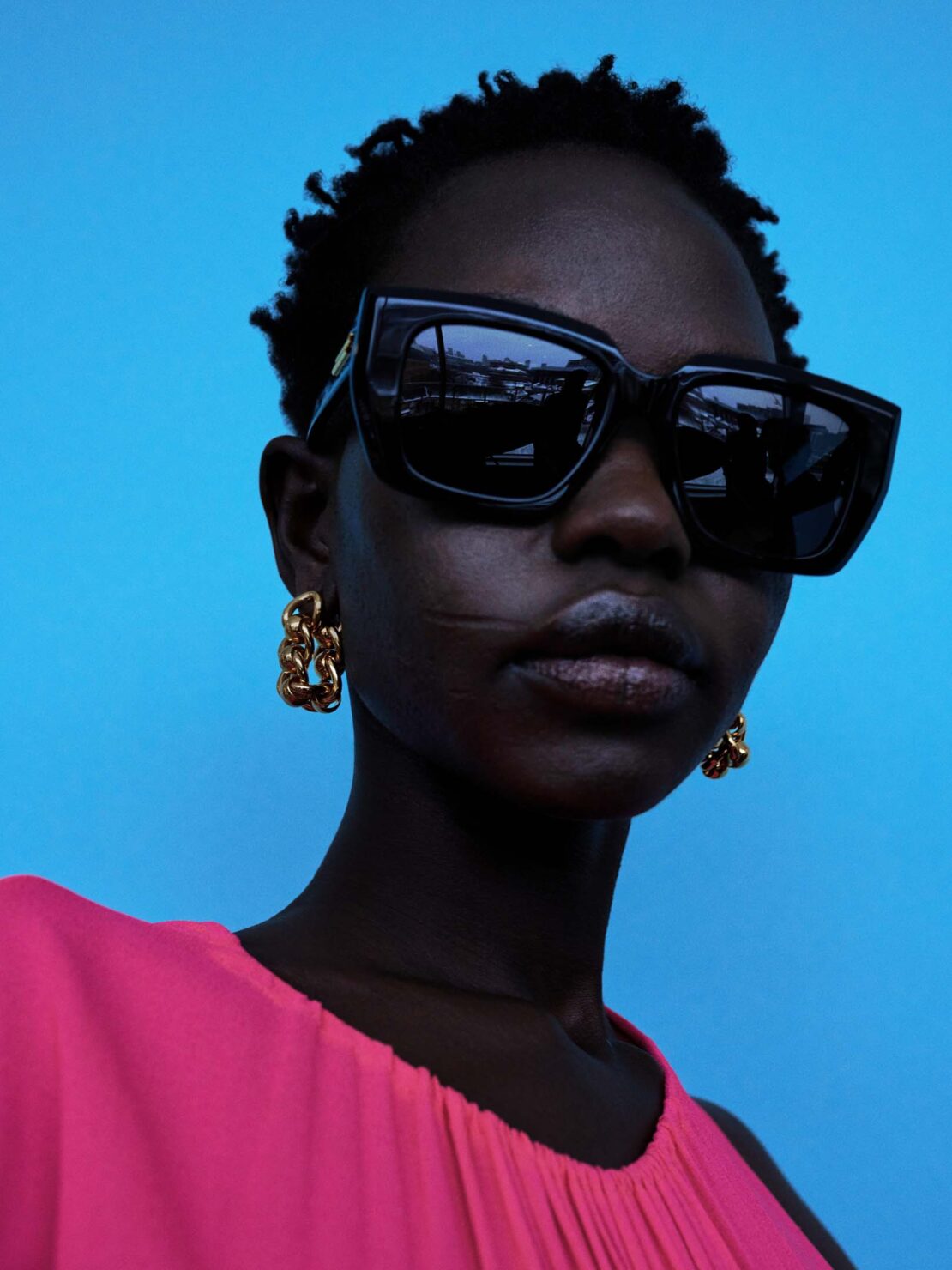 Close-up portrait of a Black woman wearing black sunglasses, gold chain earrings, and a pink top against a bright blue background.