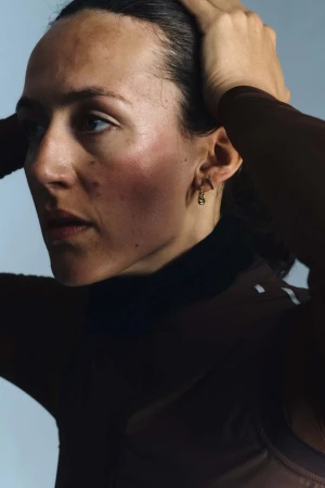 Close-up portrait of a woman with brown hair holding her hands to her head against a plain light blue background.