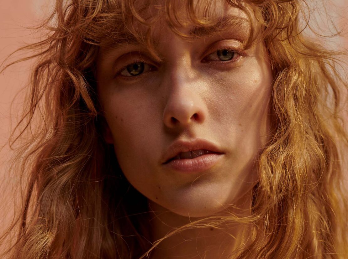 Close-up portrait of a woman with long curly hair and light eyes, captured in warm lighting by photographer Ekua King.