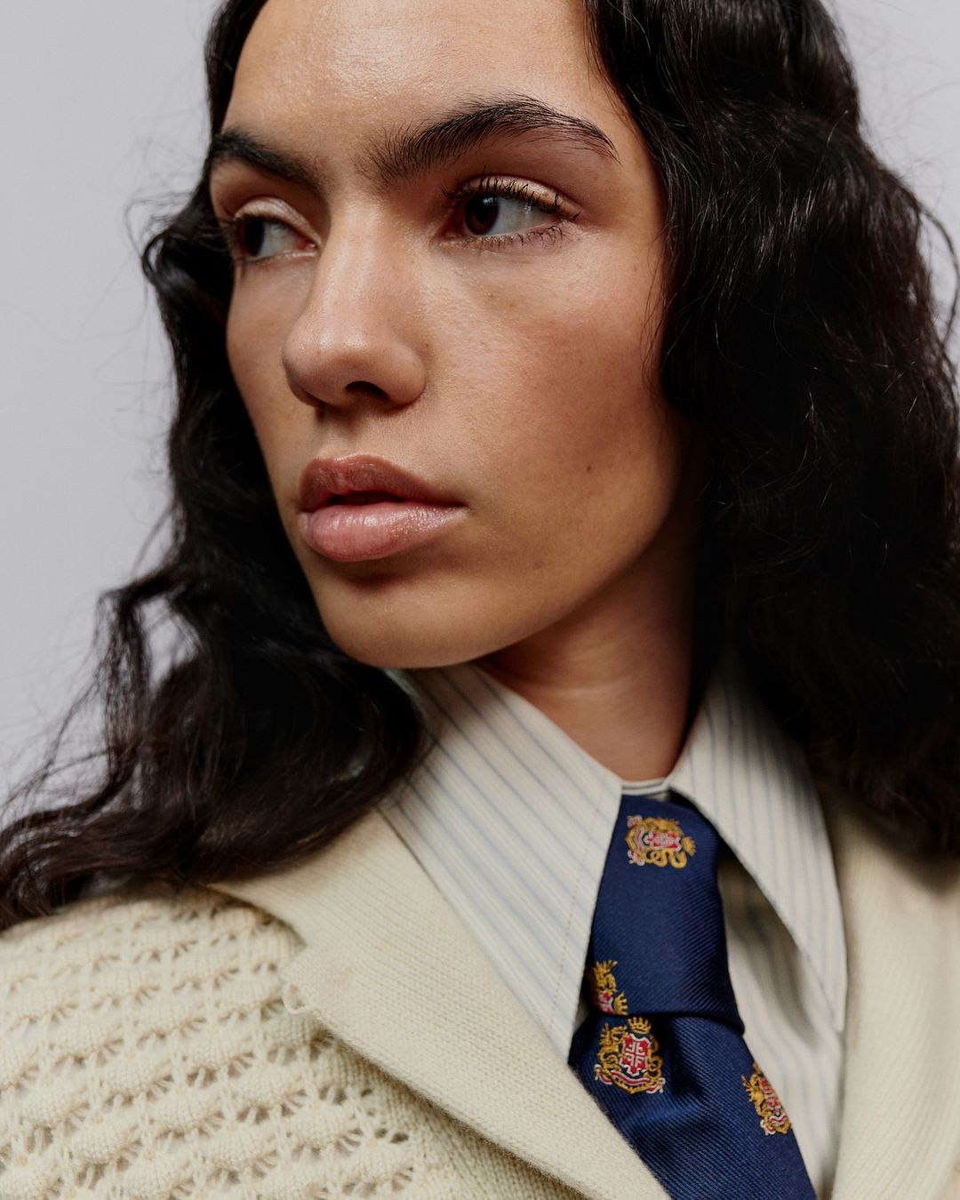 Close-up portrait of a woman with dark wavy hair wearing a patterned blue tie, striped shirt, and cream knit cardigan against a neutral wall.