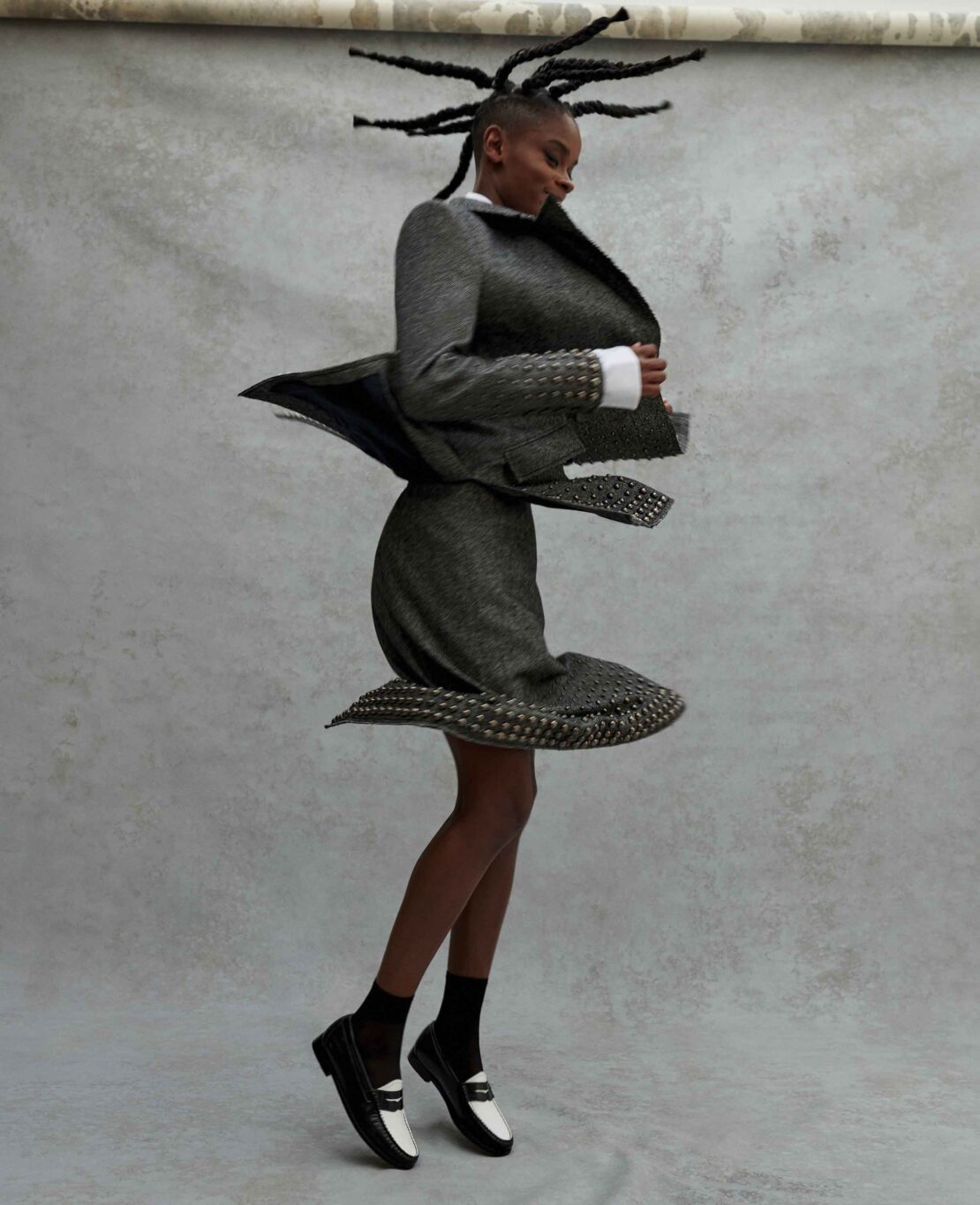 Fashion portrait of a Black woman in a gray suit with textured details, paired with black and white loafers, against a mottled backdrop.