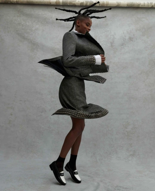 Fashion portrait of a Black woman in a gray suit with textured details, paired with black and white loafers, against a mottled backdrop.