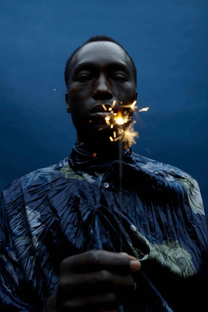 Portrait of dark-skinned man holding sparkler in front of his face, wearing blue patterned shirt, set against a dark blue background.