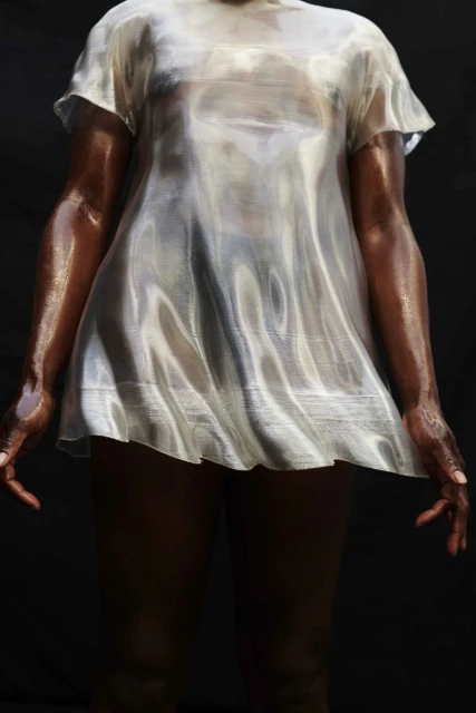 Dark-skinned adult wearing a translucent white short dress, dark background, arms at sides with hands slightly extended, studio lighting.