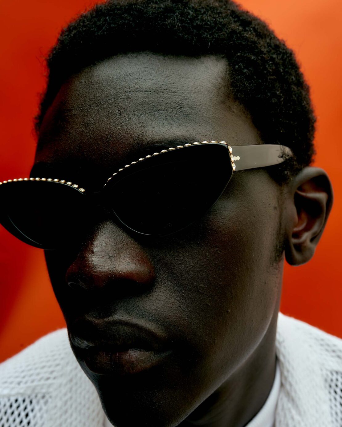Close-up portrait of an African American male model wearing black sunglasses with light trim, set against a vibrant orange backdrop.