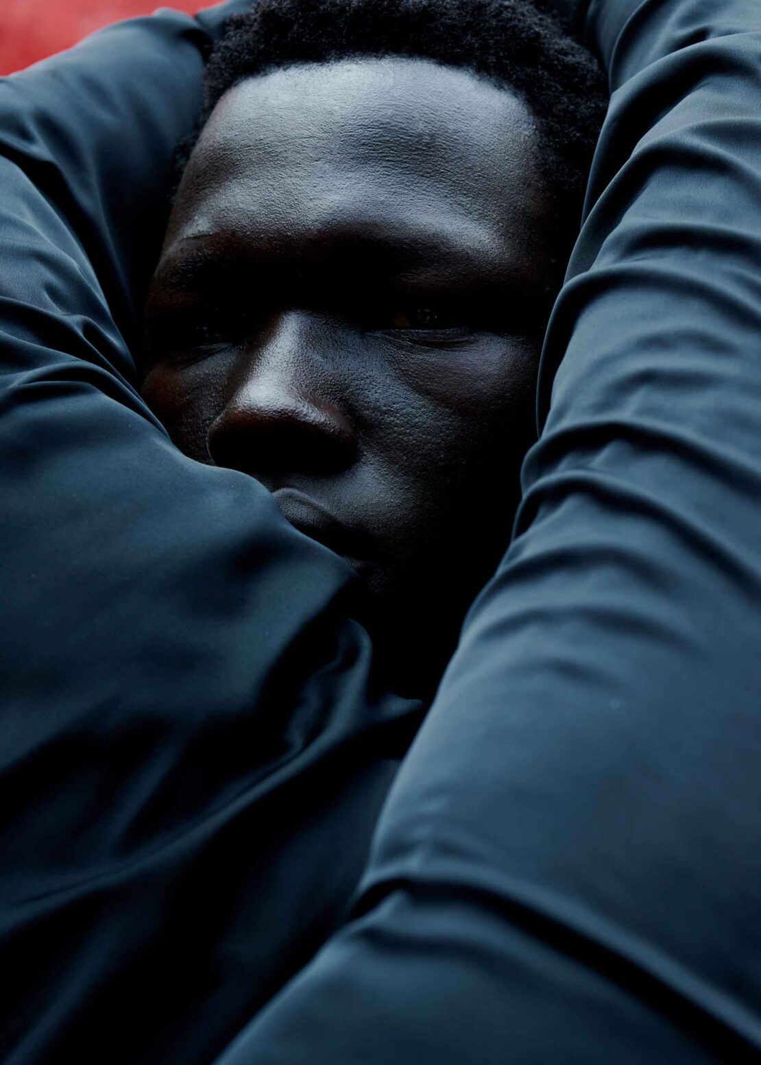 Close-up portrait of a Black male model, shown with dark hair, embracing himself with a textured black puffer jacket in the studio.