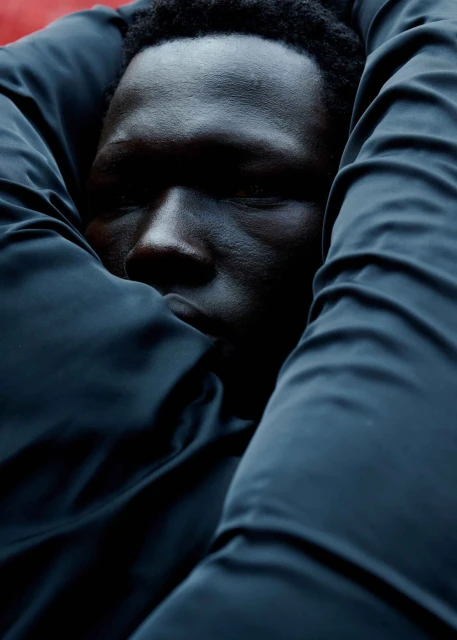 Close-up portrait of a Black male model, shown with dark hair, embracing himself with a textured black puffer jacket in the studio.