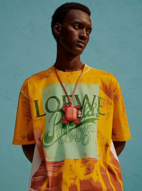 Dark-skinned male model wears a Loewe T-shirt and a turtle-shaped orange Airpod case on a strap against a blue background.
