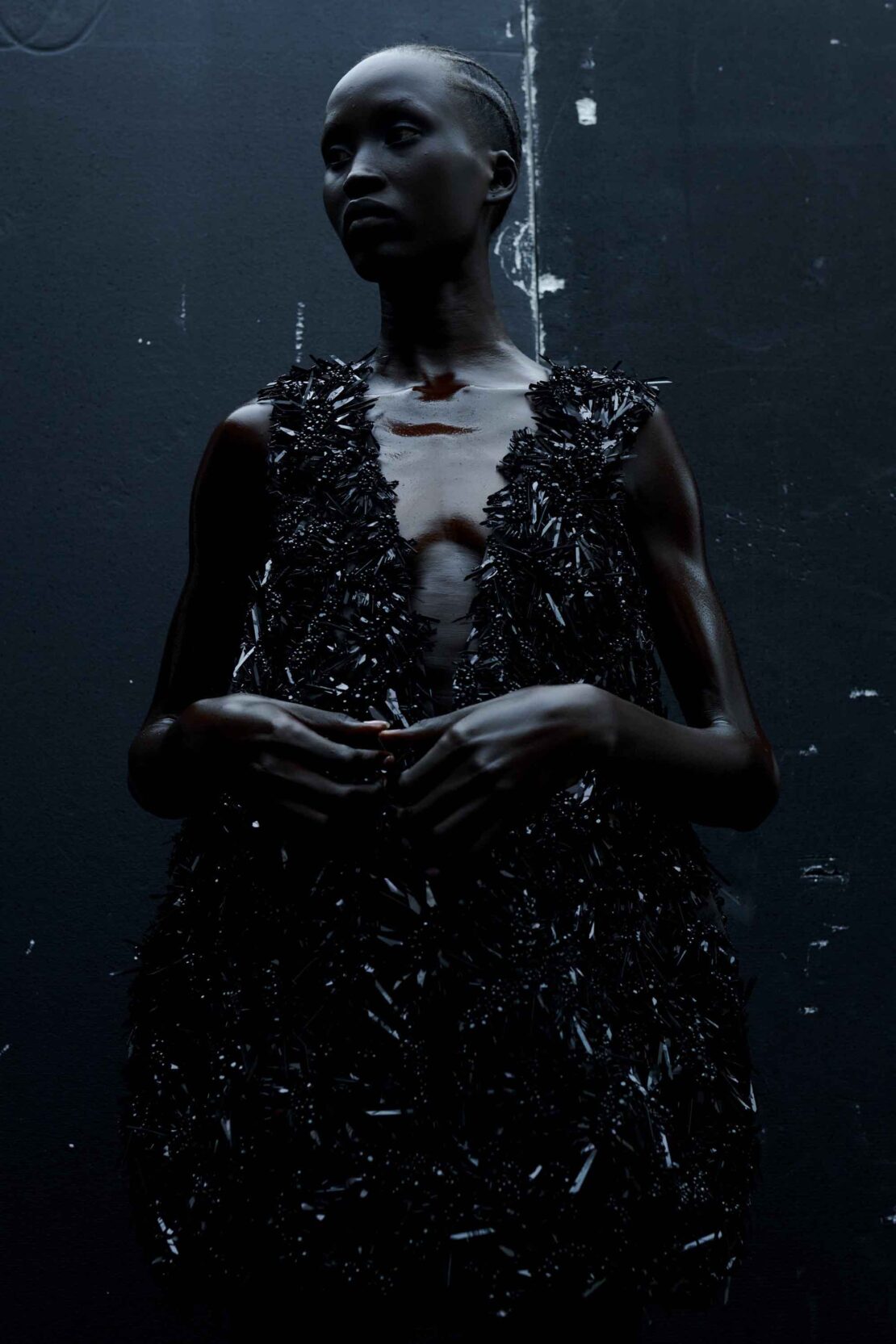 Portrait of a black woman wearing a textured black dress against a dark textured wall by photographer Ekua King