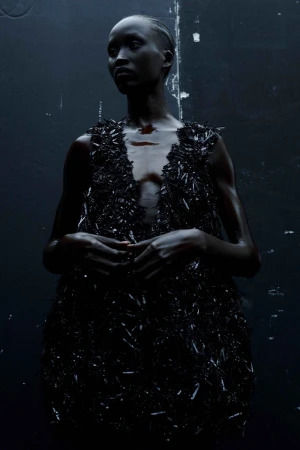 Portrait of a black woman wearing a textured black dress against a dark textured wall by photographer Ekua King