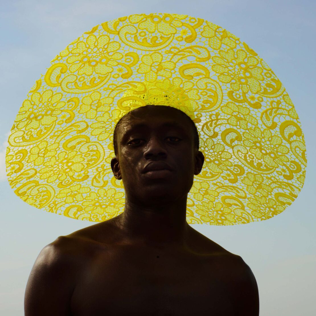 Close-up portrait of a topless black man wearing a large, yellow, lace-patterned hat against a bright, clear sky background.