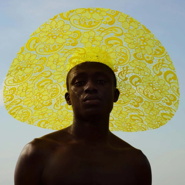Close-up portrait of a topless black man wearing a large, yellow, lace-patterned hat against a bright, clear sky background.
