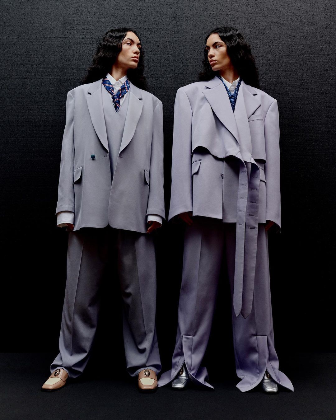 Two people stand side by side in a studio wearing oversized lavender suits, patterned scarves, and matching shoes against a dark backdrop.
