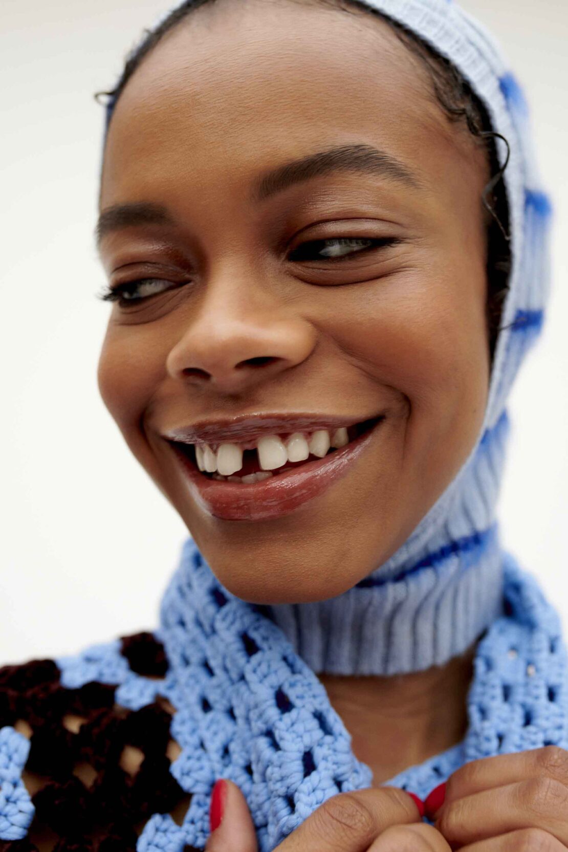 Close-up portrait of a smiling African-American woman in a blue knitted balaclava and crochet top, looking off to the side with a cheerful expression.