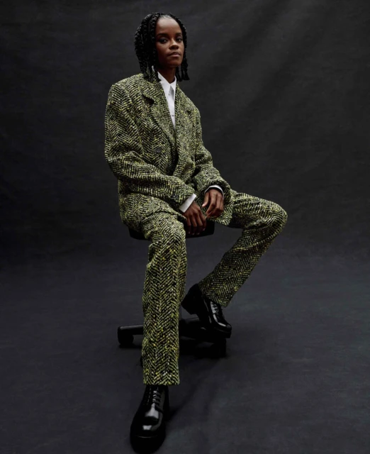 A seated woman in a green and yellow patterned suit poses in front of a dark background, wearing black shoes and a white shirt.